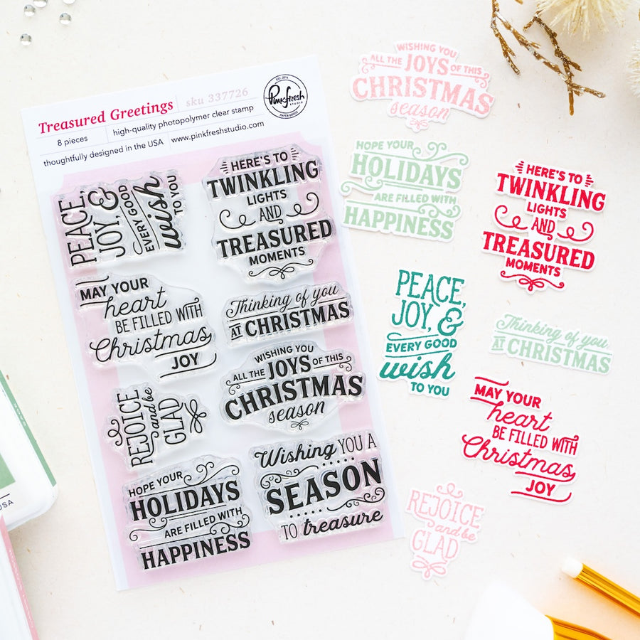 A sheet of clear holiday-themed stamp sentiments displayed on a white background, with multiple color variations of the same phrases printed beside it. The sheet reads 'Treasured Greetings' by Pinkfresh Studio and shows festive sayings like 'Peace and joy in your heart,' 'Wishing you a joyful Christmas,' and 'May your holidays be filled with Christmas joy.
