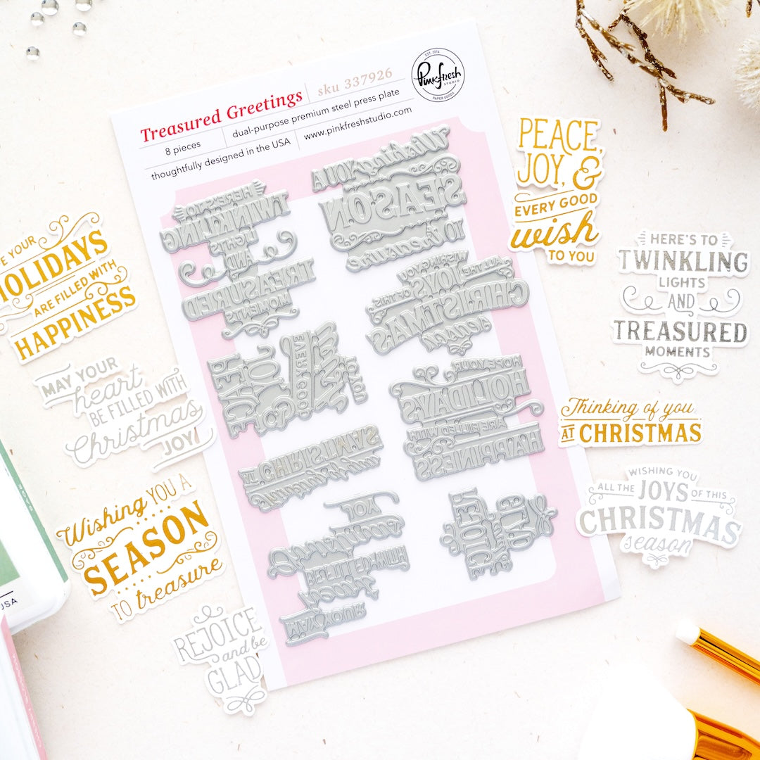 A flat-lay product photo of press plates called 'Treasured Greetings' by Pink Pinkfresh Studio. The product consists of a sheet of silver metal press plates on a light pink backing card, surrounded by sample stamp impressions in gold and black ink showing holiday greetings like 'Peace, joy, and every good wish to you' and 'Thinking of you at Christmas'.