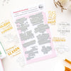 A flat-lay product photo of press plates called 'Treasured Greetings' by Pink Pinkfresh Studio. The product consists of a sheet of silver metal press plates on a light pink backing card, surrounded by sample stamp impressions in gold and black ink showing holiday greetings like 'Peace, joy, and every good wish to you' and 'Thinking of you at Christmas'.