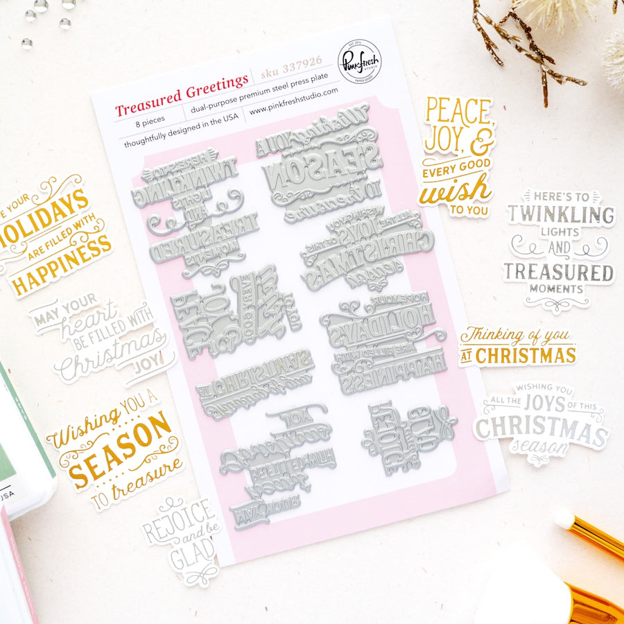 A flat-lay product photo of press plates called 'Treasured Greetings' by Pink Pinkfresh Studio. The product consists of a sheet of silver metal press plates on a light pink backing card, surrounded by sample stamp impressions in gold and black ink showing holiday greetings like 'Peace, joy, and every good wish to you' and 'Thinking of you at Christmas'.