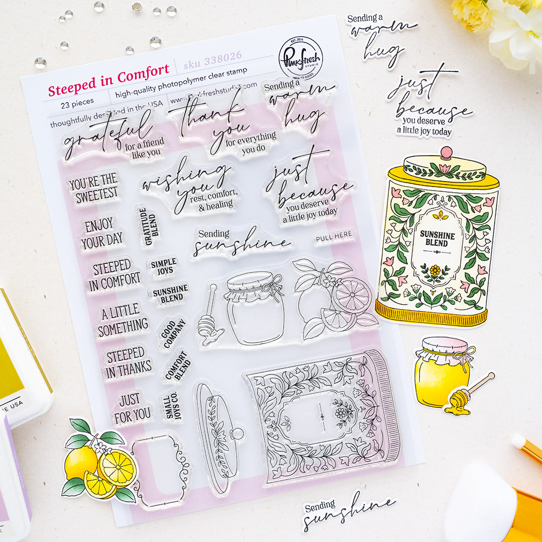 A clear stamp sheet from Pinkfresh Studio featuring cozy, warm-toned phrases like 'thank you', 'thank God', and 'just because', shown on a styled flat-lay with lemon illustrations, a honey jar, and a yellow stamp base on a white background.