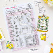 A clear stamp sheet from Pinkfresh Studio featuring cozy, warm-toned phrases like 'thank you', 'thank God', and 'just because', shown on a styled flat-lay with lemon illustrations, a honey jar, and a yellow stamp base on a white background.