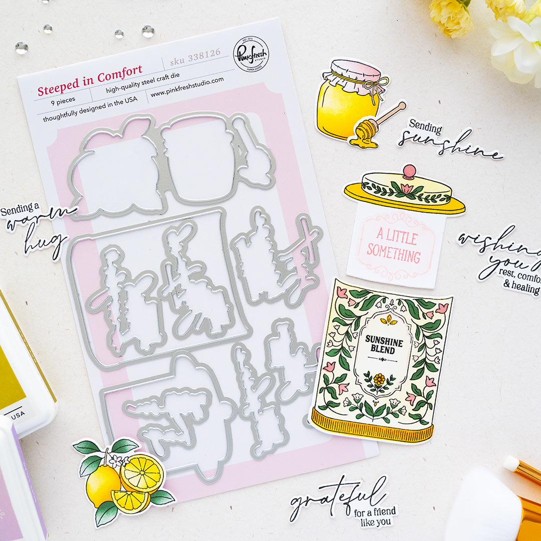 A flat-lay product photo of a craft die set and coordinating stamped/printed designs on a light surface. The main sold item appears to be the die set sheet labeled 'Steeped in Comfort' by Pinkfresh Studio with 5 pieces, shown alongside example stamped elements including honey jar, lemon, and a card design.