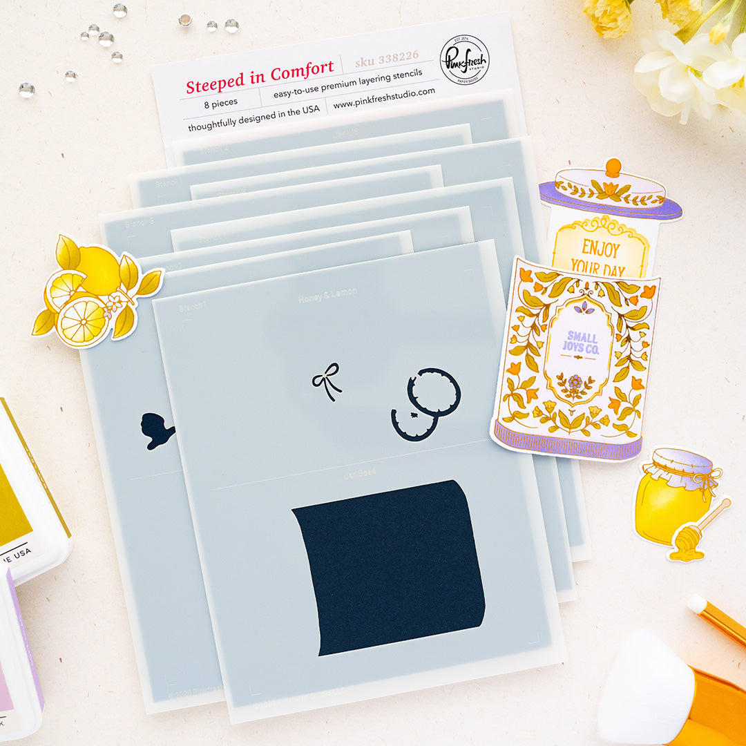 A styled flat-lay of a Pinkfresh Studio stencils set called 'Steeped in Comfort' black inked stamp impressions, surrounded by decorative stickers and branding cards on a white background.