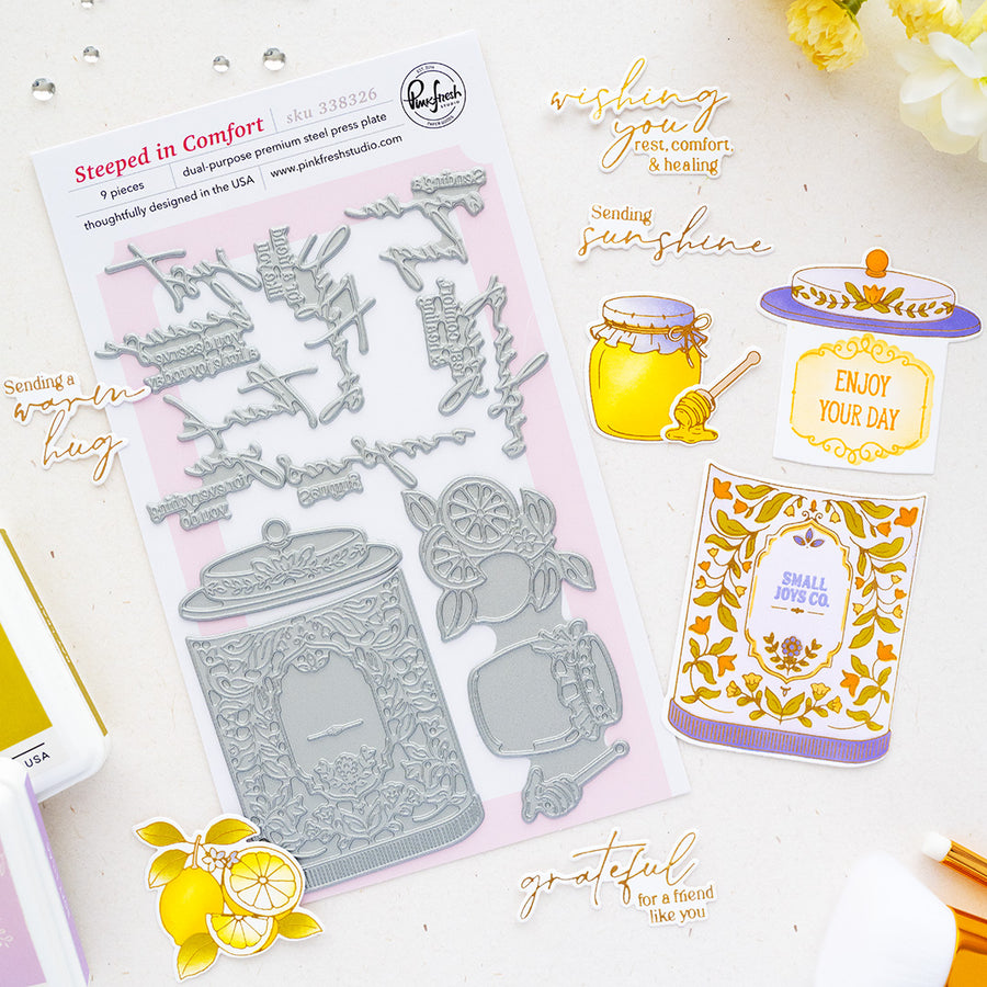 A flat-lay product photo of press plates called 'Steepled in Comfort' by Pinkfresh Studio. The kit contains a 9-piece embossed steel punch set with a clear plastic sheet showing the metal dies, alongside several example stamped cards and embellishments on a white background with decorative gold foil text and yellow flowers.