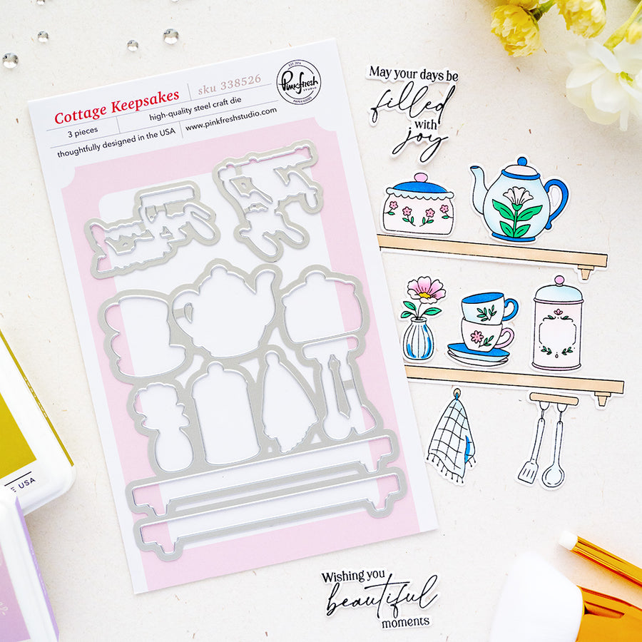 A flat-lay product photo of a steel cut die set by Pinkfresh Studio called 'Cottage Keepsakes', shown next to a stamped example sheet on a light beige background with decorative yellow flowers and craft tools.