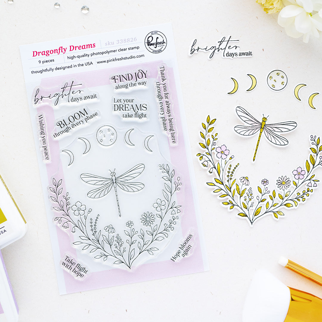 A clear stamp sheet labeled 'Dragonfly Dreams' by Pinkfresh Studio, shown on a white surface beside matching stamped artwork featuring a dragonfly, flowers, and moon phases.