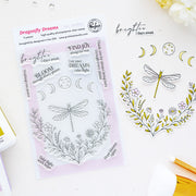 A clear stamp sheet labeled 'Dragonfly Dreams' by Pinkfresh Studio, shown on a white surface beside matching stamped artwork featuring a dragonfly, flowers, and moon phases.