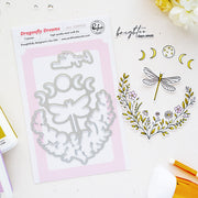 Dragonfly Dreams steel cut die set by Pinkfresh Studio, shown in its packaging alongside sample stamped and cut designs featuring a dragonfly, flowers, and crescent moons on a light background.