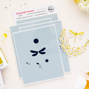 Dragonfly Dreams 4-piece premium layered stencil set by Pinkfresh Studio, displayed on a light beige surface with craft props including a gold dragonfly die-cut sticker and a white card backing.