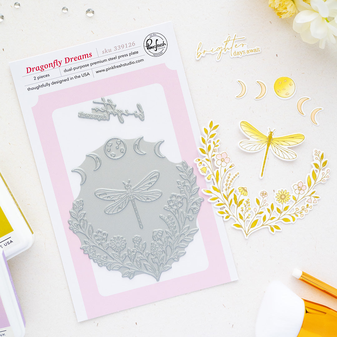 Dragonfly Dreams double-purpose steel press plate by Pinkfresh Studio shown on a white background with a pink card insert, alongside stamped and cut decorative elements including a dragonfly, floral wreath, crescent moons, and a gold pen.