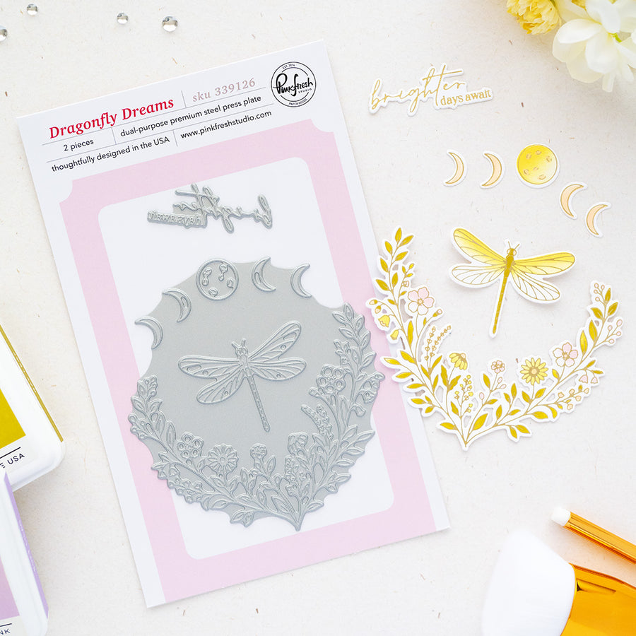 Dragonfly Dreams double-purpose steel press plate by Pinkfresh Studio shown on a white background with a pink card insert, alongside stamped and cut decorative elements including a dragonfly, floral wreath, crescent moons, and a gold pen.