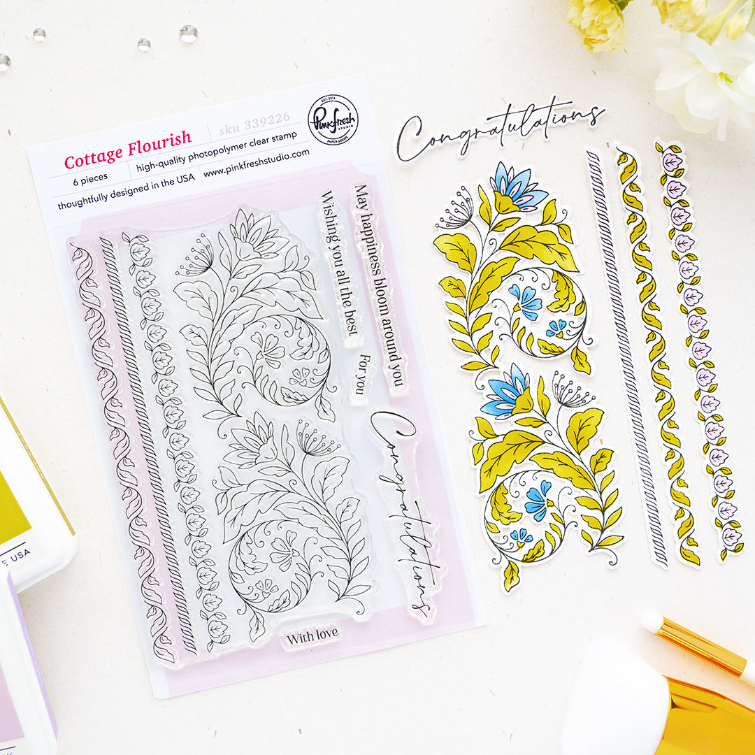 A flat-lay product photo of a clear stamp set and matching decorative stamp designs from Pinkfresh Studio called Cottage Flourish. The set includes a package of six clear stamps with floral and scroll designs, and six matching colored stamp designs in yellow, blue, and white, arranged on a white background with a gold pen and flowers as props.