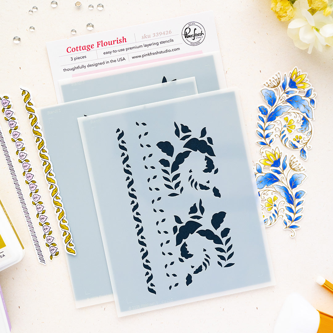 Three Cottage Flourish layering stencils by Pinkfresh Studio with floral and scroll cutouts, displayed on a light beige surface with decorative blue and gold stamped sheets and a brand card.