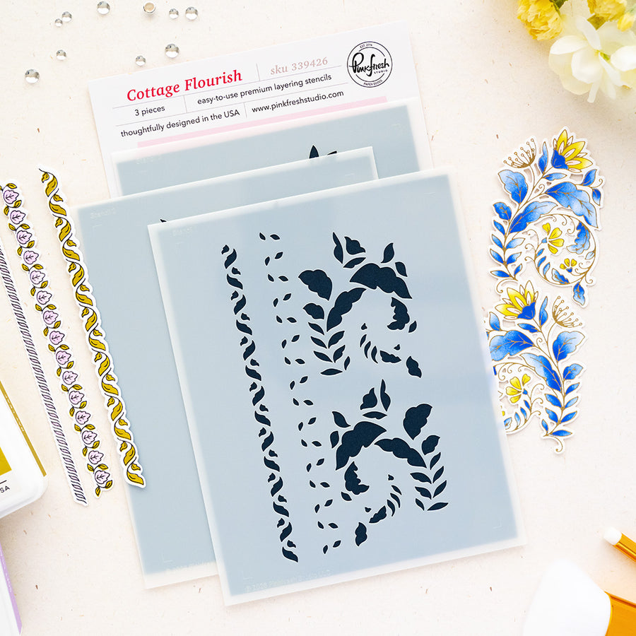 Three Cottage Flourish layering stencils by Pinkfresh Studio with floral and scroll cutouts, displayed on a light beige surface with decorative blue and gold stamped sheets and a brand card.
