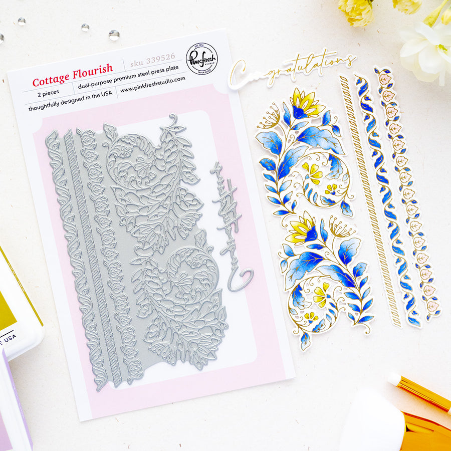 A flat-lay product photo of a stamping plate set by Pinkfresh Studio, featuring the 'Cottage Flourish' design. The left side shows the packaged steel stamp plate in its pink and white cardboard sleeve, while the right side displays the stamped floral and scroll designs in blue, gold, and white ink.