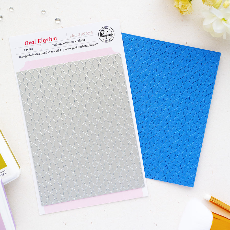 A flat-lay product photo of a pink and white paper insert for an oval mesh craft die, next to a blue textured sheet showing the die's imprint. The insert features the brand logo for 'Pinkfresh Studio' and the product name 'Oval Rhythm'.