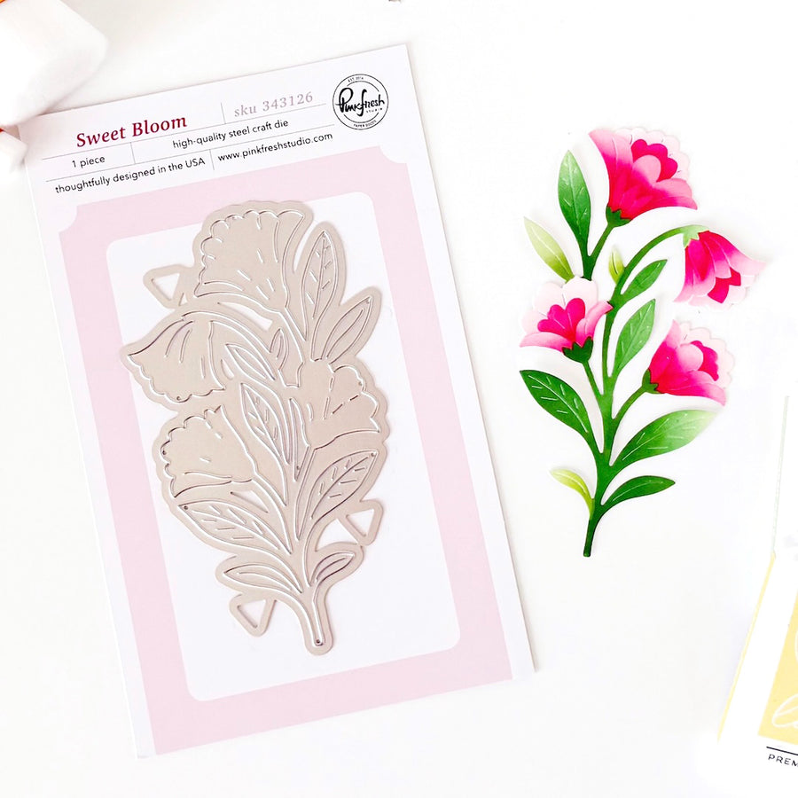 A packaged floral die-cut metal stamp set by Sweet Bloom, shown on a white background with a matching pink and green floral illustration beside it. The package reads Sweet Bloom and shows a single leafy flower die design. Pinkfresh Studio - Sweet Bloom Die