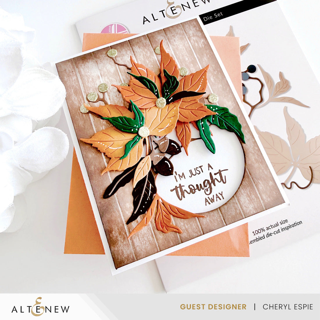 A handmade layered die-cut greeting card featuring autumn leaves in orange, green, and black over a wood-grain background, with a white oval containing the phrase 'I'm just a thought away.' The card is styled on top of other cardstock and packaging from ALTENEW, with a white flower prop on the left.