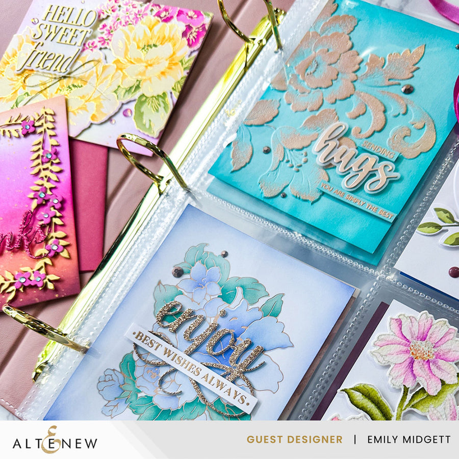 A close-up of a clear plastic ring binder holding handmade greeting cards with floral and decorative designs. Visible cards include turquoise, white, pink, and yellow floral layouts with metallic foil details.