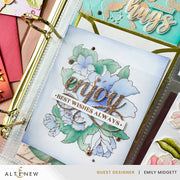 A close-up of a handmade greeting card featuring watercolor-style blue and green flowers, the word 'enjoy' in glittery gold script, and a white banner reading 'BEST WISHES ALWAYS.' The card is displayed inside a clear ring binder with gold rings, surrounded by other colorful cards and embellishments.