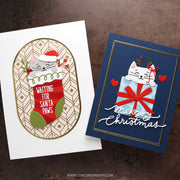 Two handmade Christmas greeting cards displayed on a dark brown textured surface. The left card is white with an oval design featuring a gray cat in a Santa hat peeking out of a red stocking that says 'WAITING FOR SANTA Paws'.