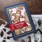 Handmade layered greeting card featuring gingerbread cookies on a pink background, a navy blue base, gold scalloped inner border, and a red ribbon banner reading 'Season's Eatings'. Card is styled on a brown wood surface with white paper cutouts and clear stamp sheets nearby.