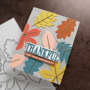 A handmade layered card featuring autumn leaf shapes in yellow, orange, blue, and pink on a white polka-dot background, with a central die-cut banner reading “THANKFUL FOR YOUR GENEROSITY.” A metal die cut file is partially visible underneath on a brown surface.