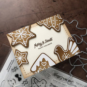 A handmade Christmas card with gingerbread cookie illustrations and the text 'Merry & Sweet Happy Holidays,' shown on a wooden surface with clear stamp sheets and metal die cutters nearby.