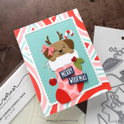 A handmade Christmas card featuring a cute cartoon reindeer tucked inside a pink stocking with 'MERRY WOOFMAS' text, shown alongside crafting tools and a stenciled paper package on a brown textured surface.
