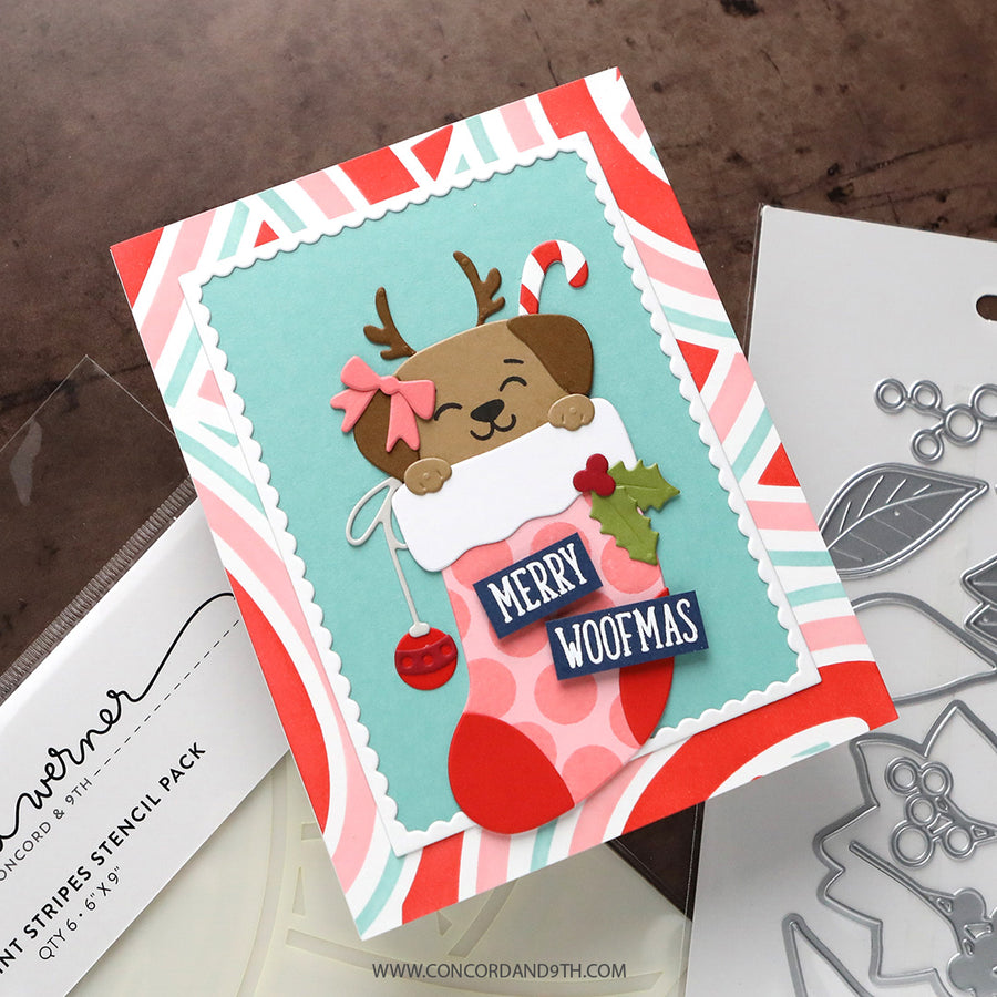 A handmade Christmas card featuring a cute cartoon reindeer tucked inside a pink stocking with 'MERRY WOOFMAS' text, shown alongside crafting tools and a stenciled paper package on a brown textured surface.