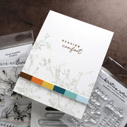 A handmade greeting card with the words 'Sending comfort' in brown script, decorated with delicate sage green botanical line art and a colorful paper strip across the bottom. The card is styled on top of clear stamp sheets on a dark brown textured surface.