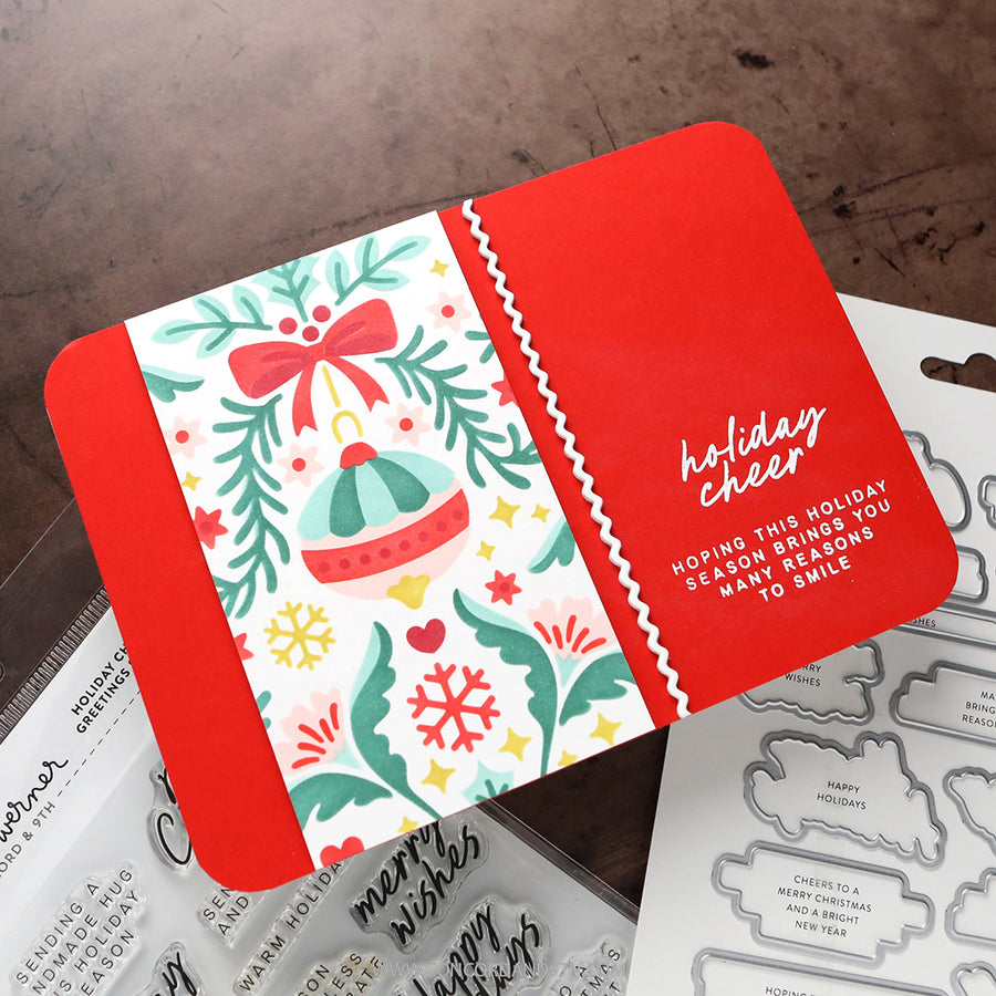 A red and white holiday-themed card or packaging piece with a colorful Christmas ornament illustration and the words 'holiday cheer' and 'hoping this holiday season brings you many reasons to smile,' photographed on a dark wood surface with craft dies and clear stamps partially visible around it.