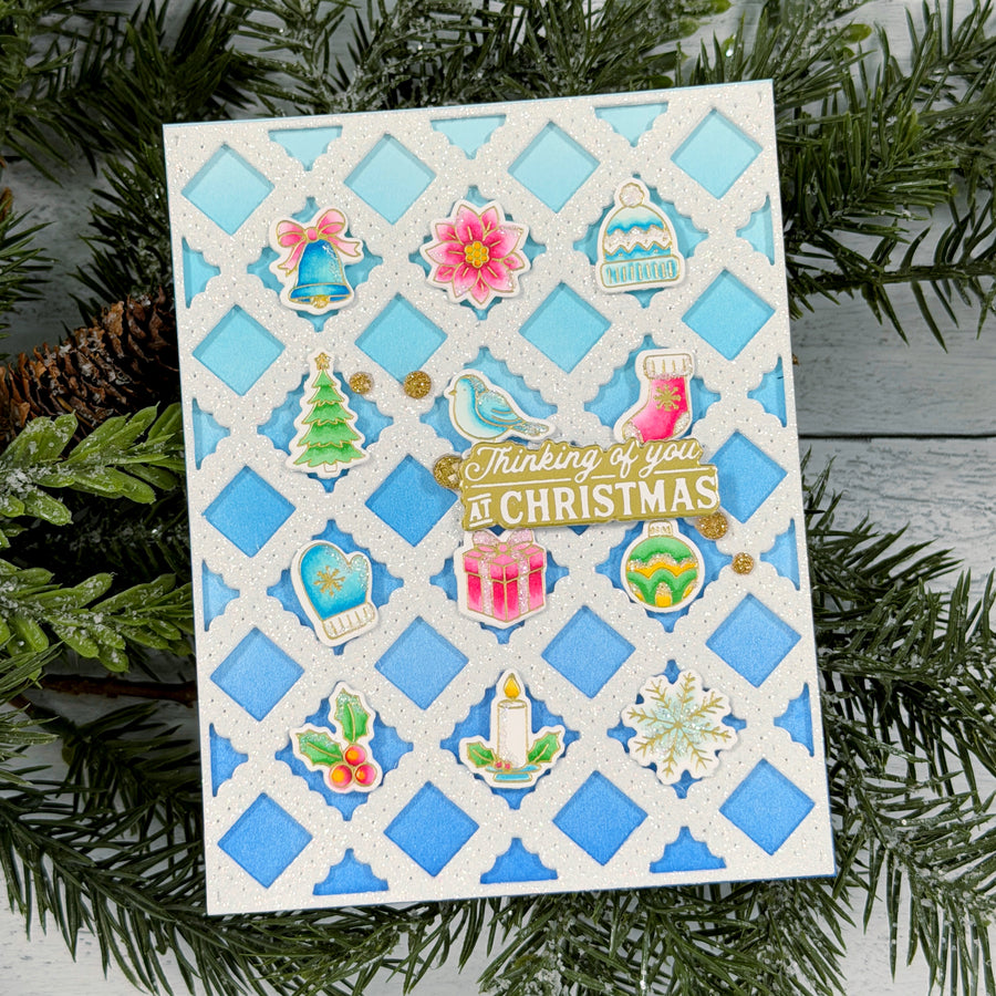 A handmade Christmas greeting card with a white trellis pattern over light blue diamonds, decorated with colorful holiday stickers and a central die-cut phrase reading 'Thinking of you AT CHRISTMAS.' The card is photographed among green pine branches on a gray wood surface.