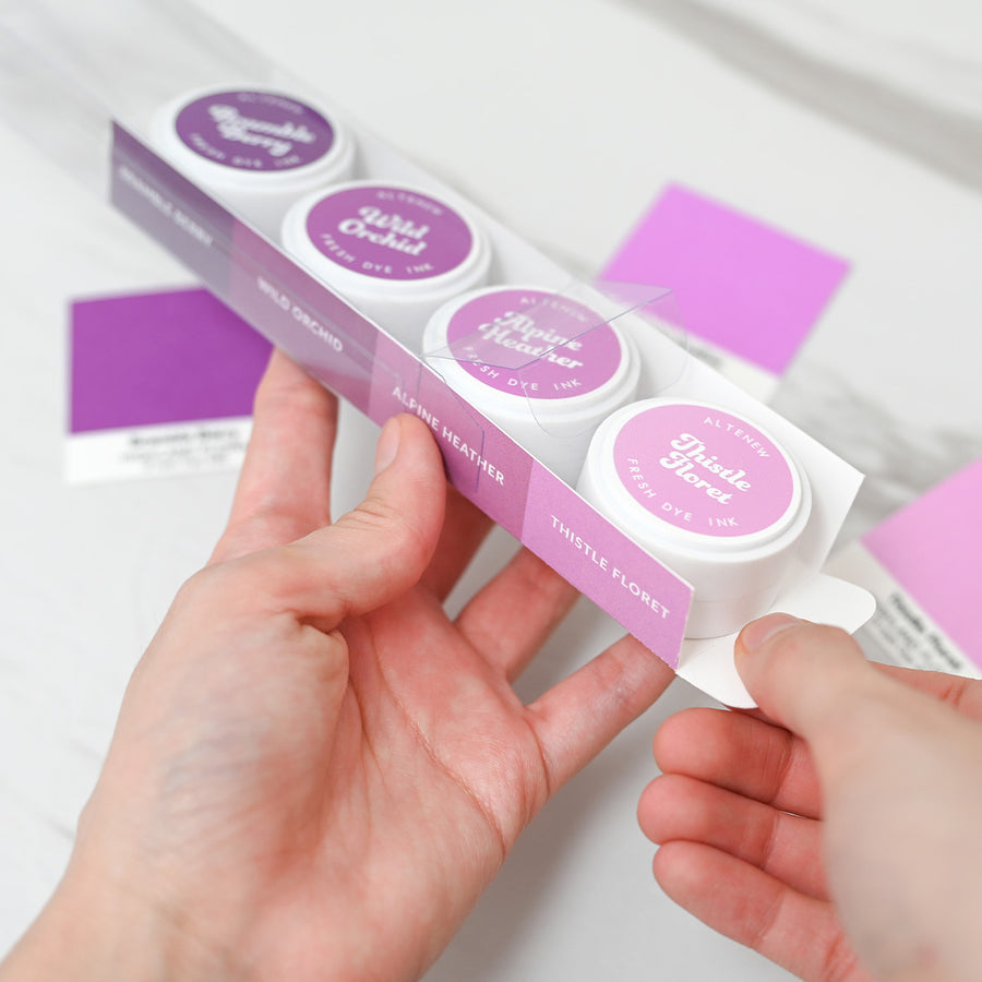 A person's hands holding a white and pink cardboard box containing three small round jars of whipped body butter. The jars have purple lids with white text reading 'Alpine Orchid' and 'Thistle Floret'. Altenew Highland Flora Fresh Dye Ink Mini Pods
