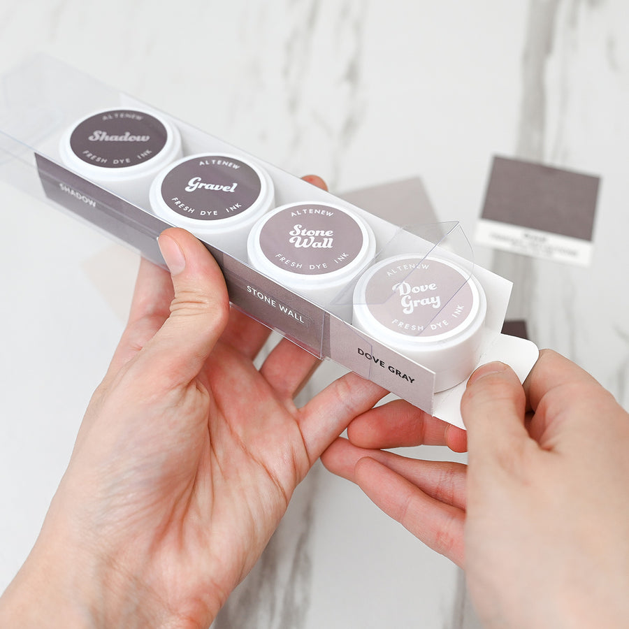 A person's hands holding a clear plastic sleeve containing four small round cosmetic pots labeled Shadow, Gravel, Stone Wall, and Dove Gray, against a light marble background. Altenew Tranquil Reflections Fresh Dye Ink Mini Pods