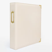 An ivory-colored faux leather ring binder with gold-tone metal accents, including a decorative side handle and corner protectors, shown on a clean white background. 