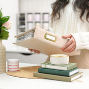 A person in a cream cable-knit sweater holds an ivory faux-leather binder with a gold-tone metal nameplate. The scene is set in a bright, modern room with a styled desk featuring stacked green and beige books, a small white candle with gold trim, and a pink rose in a textured vase. 