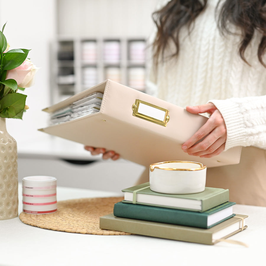 A person in a cream cable-knit sweater holds an ivory faux-leather binder with a gold-tone metal nameplate. The scene is set in a bright, modern room with a styled desk featuring stacked green and beige books, a small white candle with gold trim, and a pink rose in a textured vase. 
