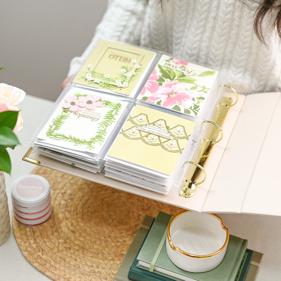 A person holds an open ring binder filled with decorative paper cards or stickers in pastel floral and geometric designs. The scene is styled on a tabletop with a woven placemat, a small white dish, books, and flowers, suggesting a lifestyle presentation of stationery or craft supplies. 