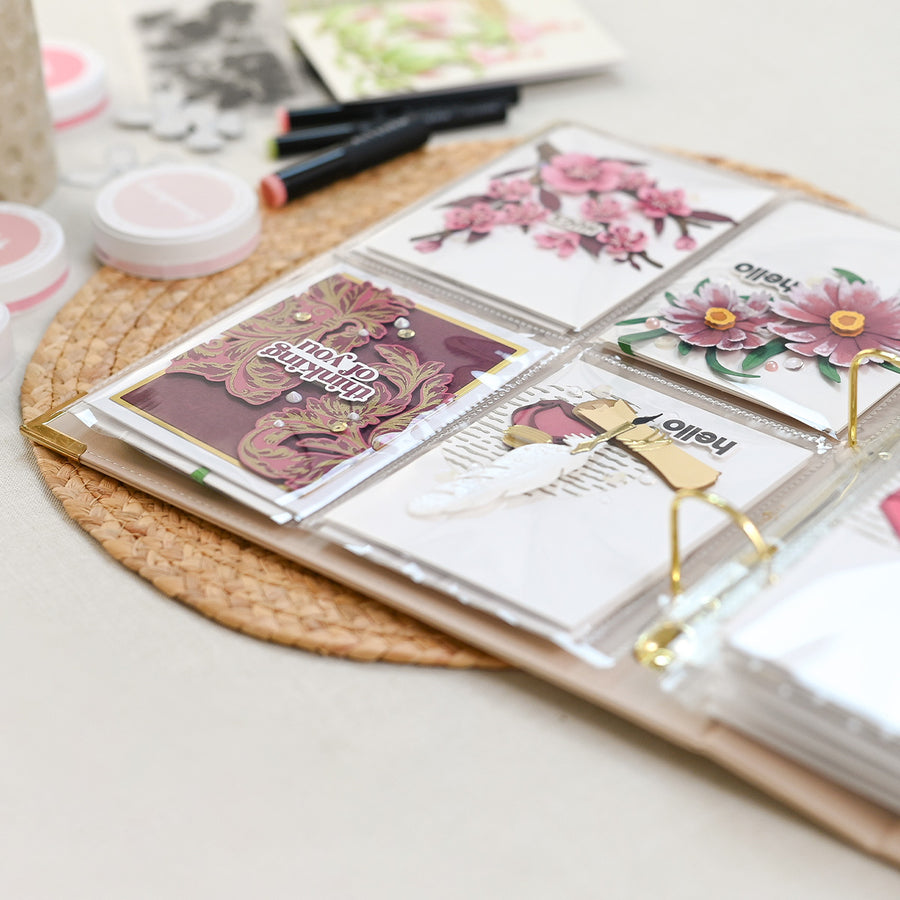 An open clear plastic binder or album with several decorated sticker or card inserts displayed in the rings, photographed on a woven round mat with makeup items blurred in the background. 