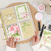 A top-down view of a person's hands holding an open ring binder filled with floral and decorative paper cards. The scene includes small cosmetic-style jars, black and pink pens, and additional card designs arranged on a woven mat and light fabric background. 