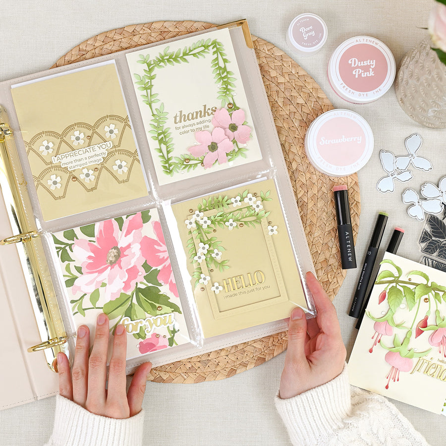 A top-down view of a person's hands holding an open ring binder filled with floral and decorative paper cards. The scene includes small cosmetic-style jars, black and pink pens, and additional card designs arranged on a woven mat and light fabric background. 