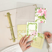 A top-down view of a clear binder with a gold ring holding handmade greeting cards. Two hands are visible holding the cards in place on a light fabric surface. 