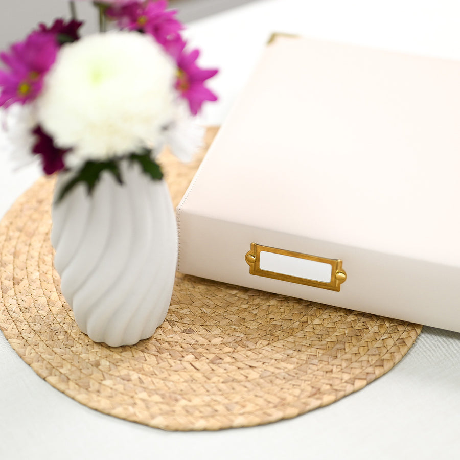 A ivory-colored paper organizer box with a gold metal label holder, styled on a woven placemat next to a white ribbed ceramic vase with white and purple flowers. 