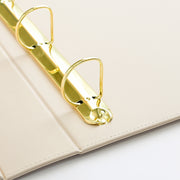Close-up of an ivory-colored faux leather ring binder with shiny gold metal rings, showing the stitched edge and hinge detail. 