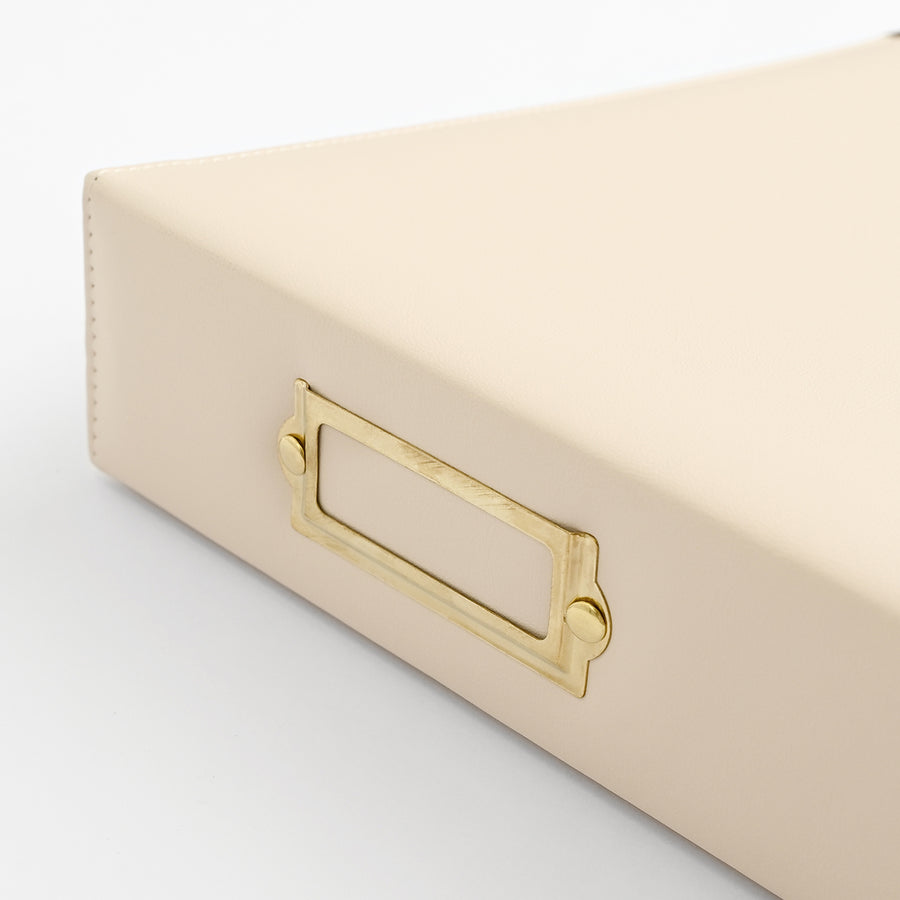 A close-up of an ivory-colored leather or faux-leather storage box with a gold metal label holder handle, set against a clean white background. 