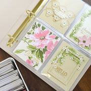A close-up of a ring-bound card album or binder open on a wooden surface, showing four decorated card inserts with floral and decorative designs in soft pink, green, cream, and gold tones. A metal tin of cards sits at the lower left. 