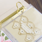 A close-up of a clear plastic binder ring holding a cream-colored card with gold scalloped outlines, white flower embellishments, and the phrase 'I APPRECIATE YOU' on a wooden surface. 