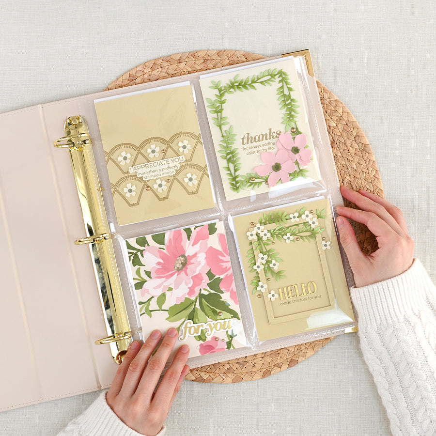 A clear four-pocket binder with a pale ivory cover and gold-tone ring hardware, shown open on a woven placemat. The pockets hold decorative cards with floral and geometric designs, and two hands are holding the binder open. 